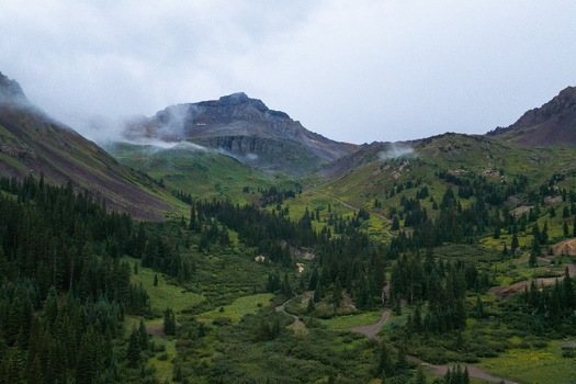 Aerial view of Mt. Sneffels and the lush green landscape of Yankee Boy Basin with pine forests winding trails and low hanging clouds in Ouray Colorado United States.