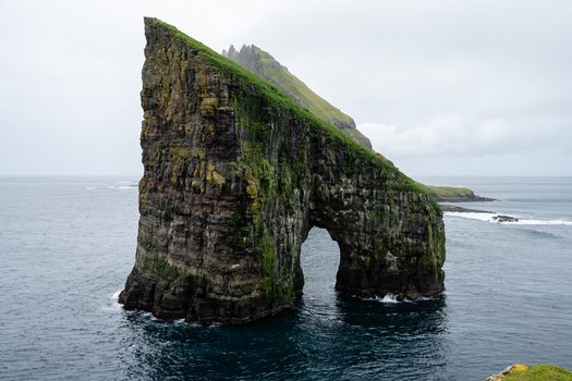 View of the dramatic Drangarnir sea stacks with a natural arch and mossy green cliffs rising from the dark ocean Faroe Islands.