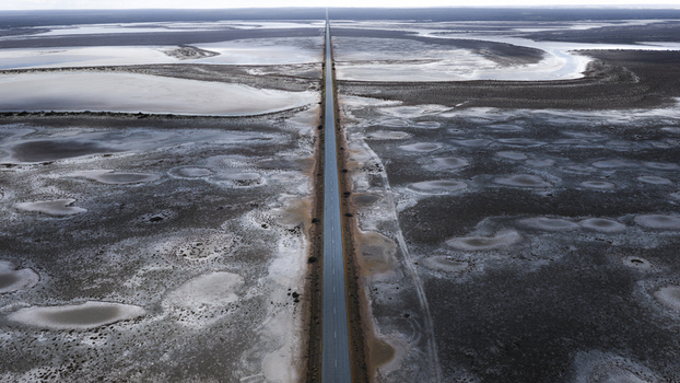 Aerial view of a long straight road cutting through a vast salt crusted landscape with shallow pools of water and dark textured terrain in Perth Western Australia Australia.