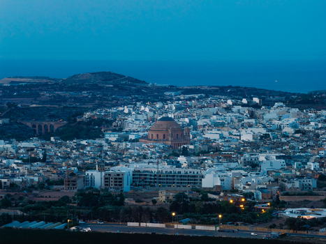 Aerial view of the Mosta Rotunda dome rising above the dense urban landscape of Mosta at dusk with surrounding limestone buildings and distant hills. Mosta Western Region Malta. Aerial view of the Mosta Rotunda dome rising above the dense urban landscape of Mosta at dusk with surrounding limestone buildings and distant hills. Mosta Western Region Malta.