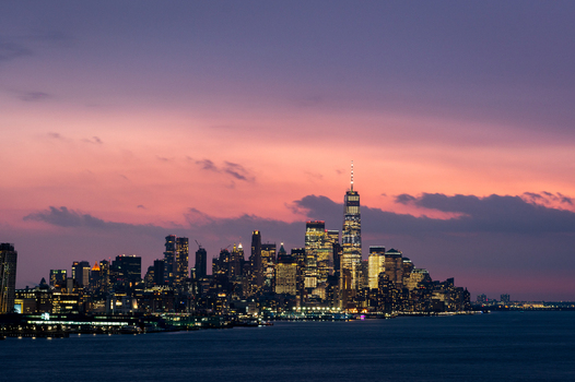 View of the New York City skyline at sunset featuring the One World Trade Center illuminated against a vibrant purple and orange sky over the Hudson River United States of America. View of the New York City skyline at sunset featuring the One World Trade Center illuminated against a vibrant purple and orange sky over the Hudson River United States of America.