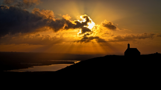 Aerial view of Chapelle Saint Michel de Brasparts silhouetted on a hill with dramatic sun rays piercing through golden clouds over a lake Saint Rivoal Bretagne France. Aerial view of Chapelle Saint Michel de Brasparts silhouetted on a hill with dramatic sun rays piercing through golden clouds over a lake Saint Rivoal Bretagne France.