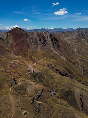 Aerial view of Vinicunca Rainbow Mountain showing colorful mineral layers and winding hiking trails with snow capped Andes peaks in the distance Quispicanchi Province Cusco Peru.
