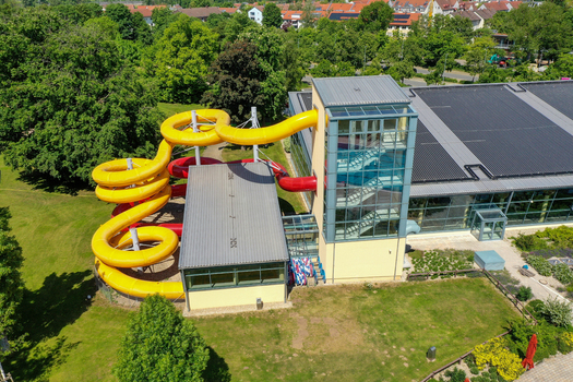 Aerial view of Konigsbad Forchheim featuring vibrant yellow and red water slides and a modern glass staircase tower surrounded by lush green trees in Forchheim Bayern Germany. Aerial view of Konigsbad Forchheim featuring vibrant yellow and red water slides and a modern glass staircase tower surrounded by lush green trees in Forchheim Bayern Germany.