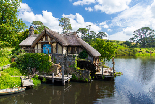 View of rustic mill house with thatched roof and blue shutters nestled by a serene lake under a bright sky Hobbiton Movie Set Waikato Region New Zealand. View of rustic mill house with thatched roof and blue shutters nestled by a serene lake under a bright sky Hobbiton Movie Set Waikato Region New Zealand.