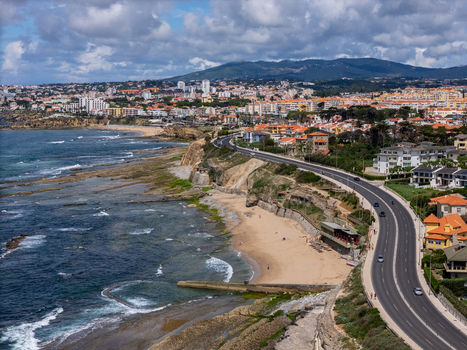 Aerial view of the Avenida Marginal coastal road winding along the rocky shoreline and sandy beach with residential buildings and hills under a cloudy sky in Parede Lisboa Portugal. Aerial view of the Avenida Marginal coastal road winding along the rocky shoreline and sandy beach with residential buildings and hills under a cloudy sky in Parede Lisboa Portugal.