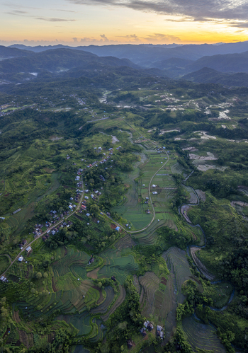 Aerial view of Bangka Kenda rice terraces surrounded by lush green hills winding roads and small village houses under a golden sunset sky in Wae Rii East Nusa Tenggara Indonesia.