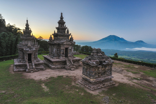 View of the ancient stone structures of Gedong Songo Temple with mountain peaks in the distance under a soft morning sky Bandungan Jawa Tengah Indonesia.