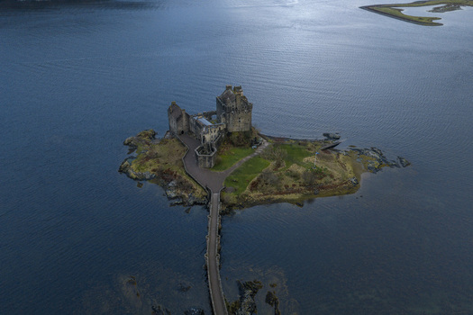 Aerial view of Eilean Donan Castle on a small tidal island connected by a stone bridge surrounded by calm dark waters in Dornie Scotland United Kingdom.