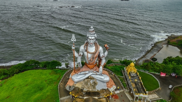 Aerial view of the massive Shiva statue at Murudeshwar Temple featuring its silver finish and trident set against the backdrop of the Arabian Sea in Murdeshwar Karnataka India.