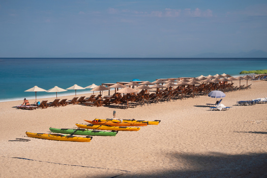 View of golden sand meets turquoise water under a bright sky with rows of beach umbrellas and colorful kayaks lining the shore Lefkada Greece. View of golden sand meets turquoise water under a bright sky with rows of beach umbrellas and colorful kayaks lining the shore Lefkada Greece.