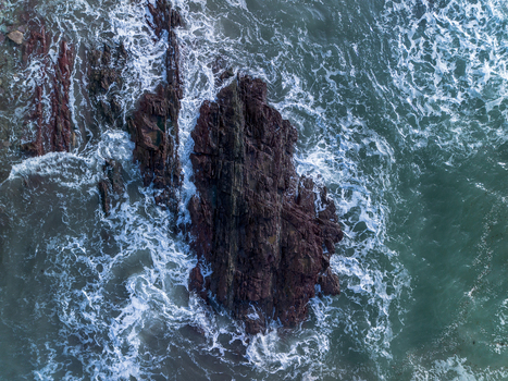 Aerial view of jagged dark rocks along the coastline with dramatic waves crashing and white sea foam in Talland Bay Cornwall United Kingdom. Aerial view of jagged dark rocks along the coastline with dramatic waves crashing and white sea foam in Talland Bay Cornwall United Kingdom.