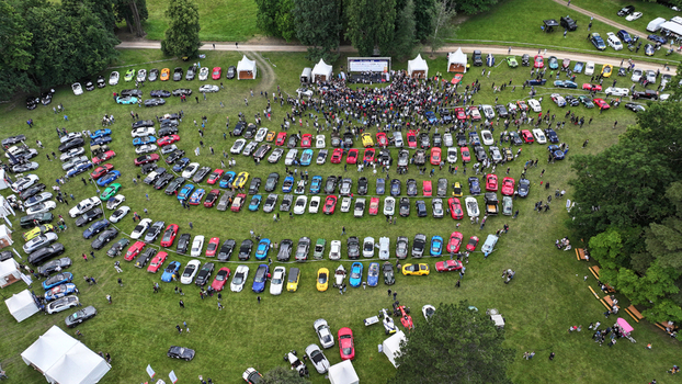 La Chapelle de Guinchay France 01 June 2024 Aerial view of a large car show with rows of colorful vehicles parked on a green field and crowds of people gathered near a stage.