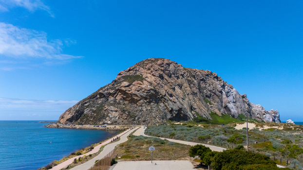 Aerial view of Morro Rock by the Pacific Ocean with a basketball court and coastal roads under a clear blue sky in Morro Bay California United States.