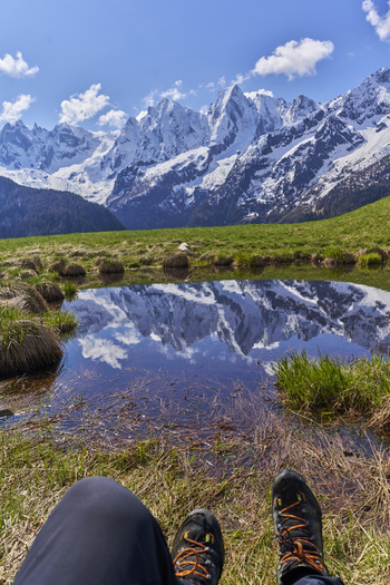 View of the Engadin valley with snow capped mountains reflecting in a small alpine pond seen from the perspective of a hiker wearing boots in a green meadow Grisons Switzerland. View of the Engadin valley with snow capped mountains reflecting in a small alpine pond seen from the perspective of a hiker wearing boots in a green meadow Grisons Switzerland.