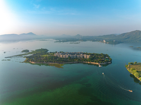 Aerial view of the historic island palace in Lake Pichola surrounded by lush greenery and calm waters with distant hills under a clear sky in Udaipur Rajasthan India. Aerial view of the historic island palace in Lake Pichola surrounded by lush greenery and calm waters with distant hills under a clear sky in Udaipur Rajasthan India.