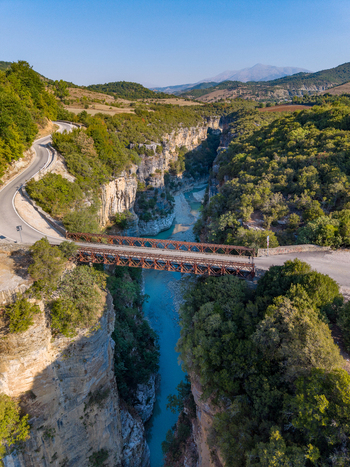 Aerial view of Osumi canyon with a metal bridge crossing the turquoise river surrounded by steep rocky cliffs lush green trees and winding roads in Dhores Berat County Albania.
