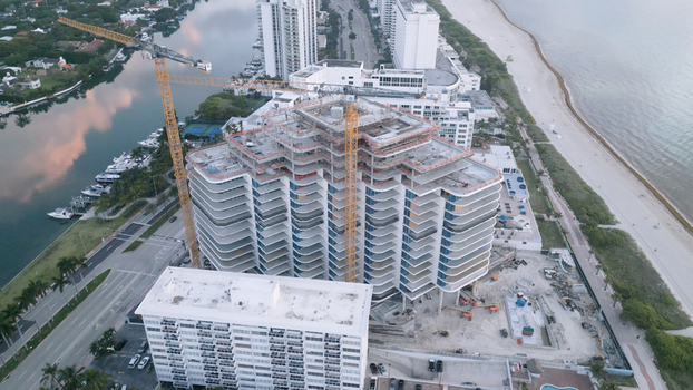 Aerial view of a high rise building under construction with two yellow tower cranes situated between a canal with docked boats and the sandy beach in Miami Beach Florida United States. Aerial view of a high rise building under construction with two yellow tower cranes situated between a canal with docked boats and the sandy beach in Miami Beach Florida United States.