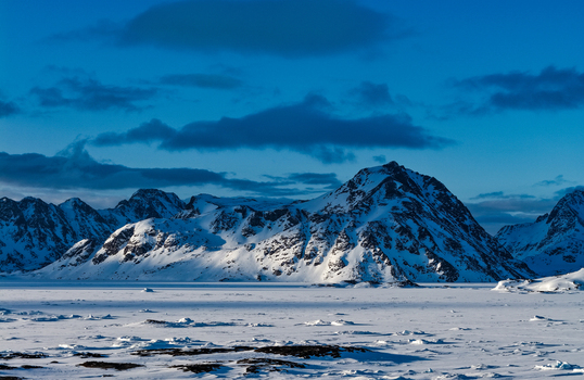 Aerial view of snow covered mountain peaks and a vast frozen arctic landscape under a deep blue sky with dark clouds in Tasiilaq Sermersooq Municipali Greenland. Aerial view of snow covered mountain peaks and a vast frozen arctic landscape under a deep blue sky with dark clouds in Tasiilaq Sermersooq Municipali Greenland.