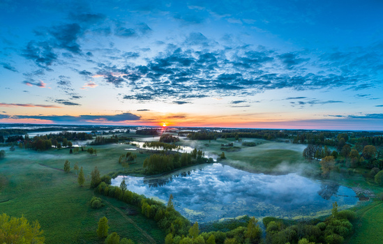 Aerial view of a serene landscape in Latvia featuring a misty lake surrounded by lush green fields and trees at sunrise under a vibrant sky. Aerial view of a serene landscape in Latvia featuring a misty lake surrounded by lush green fields and trees at sunrise under a vibrant sky.