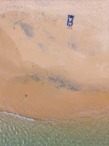 Aerial view of a person sunbathing on a blue towel on the golden sand near the clear turquoise water in Cascais Lisbon Portugal. Aerial view of a person sunbathing on a blue towel on the golden sand near the clear turquoise water in Cascais Lisbon Portugal.