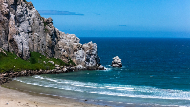 Aerial view of the massive Morro Rock volcanic plug towering over the sandy beach and blue Pacific Ocean waves under a clear sky in Morro Bay California United States.