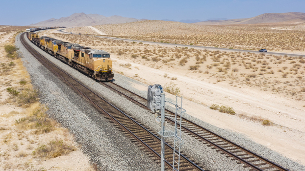 Aerial view of a freight train traveling through a vast desert landscape with mountains in the background and a nearby highway in the United States.