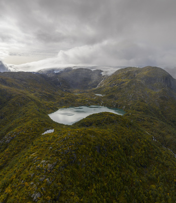 Aerial view of the Buarbreen glacier area and a turquoise mountain lake nestled between rugged rocky peaks and green slopes under a cloudy sky in Ullensvang Vestland Norway. Aerial view of the Buarbreen glacier area and a turquoise mountain lake nestled between rugged rocky peaks and green slopes under a cloudy sky in Ullensvang Vestland Norway.