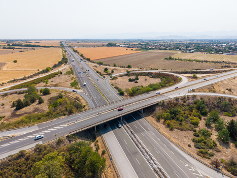 Aerial view of Trakiya Highway interchange with vehicles driving on asphalt roads surrounded by agricultural fields and rural landscape in Bulgaria. Aerial view of Trakiya Highway interchange with vehicles driving on asphalt roads surrounded by agricultural fields and rural landscape in Bulgaria.