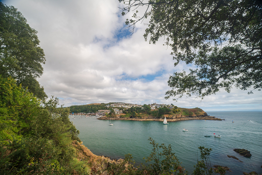 View of the Fowey River with boats sailing near the historic town of Fowey framed by lush trees and a cloudy sky in Fowey Cornwall United Kingdom. View of the Fowey River with boats sailing near the historic town of Fowey framed by lush trees and a cloudy sky in Fowey Cornwall United Kingdom.