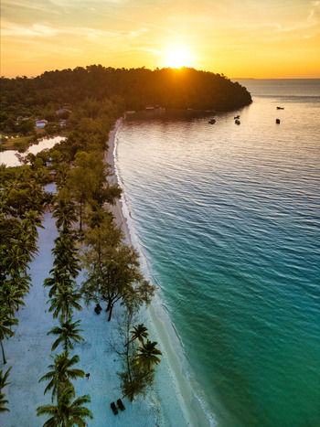 Aerial view of a sun kissed beach with turquoise waters meeting the soft sandy shore framed by lush green trees under a golden sunset Preah Sihanouk Sihanoukville Cambodia.