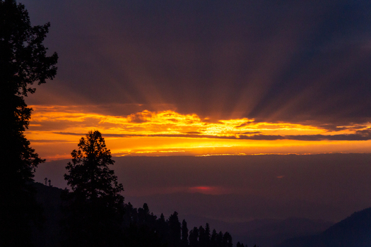 View of the sun s radiant rays pierce through dark shadowy clouds over a silhouette of trees on a hillside creating a dramatic scene Pir Chanasi Azad Kashmir Pakistan. View of the sun s radiant rays pierce through dark shadowy clouds over a silhouette of trees on a hillside creating a dramatic scene Pir Chanasi Azad Kashmir Pakistan.