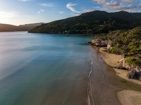 Aerial view of a serene coastal beach with calm turquoise waters golden sand and lush green hills under a soft sunset sky on Great Barrier Island New Zealand. Aerial view of a serene coastal beach with calm turquoise waters golden sand and lush green hills under a soft sunset sky on Great Barrier Island New Zealand.