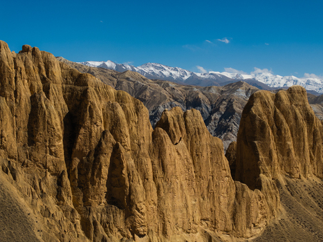 Aerial view of rugged eroded golden brown rock formations with snow capped Himalayan peaks in the background under a clear blue sky in Nepal.