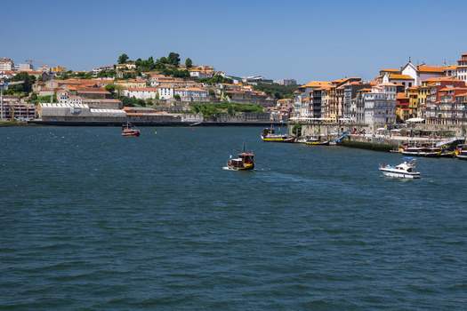Aerial view of the Douro River reflecting the bright sky as boats navigate past Ribeira s colorful buildings and terracotta roofs Porto Porto District Portugal. Aerial view of the Douro River reflecting the bright sky as boats navigate past Ribeira s colorful buildings and terracotta roofs Porto Porto District Portugal.
