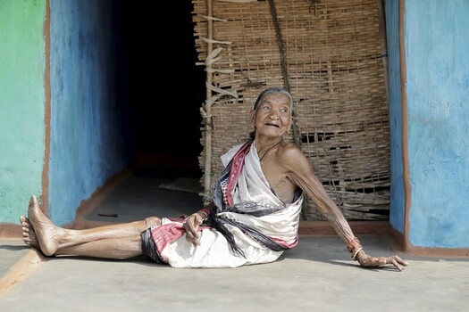 Odisha India 26 March 2026 View of an elderly woman with wrinkled skin sits relaxed on the ground against a weathered blue wall and woven door. Odisha India 26 March 2026 View of an elderly woman with wrinkled skin sits relaxed on the ground against a weathered blue wall and woven door.