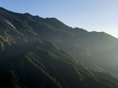 Aerial view of the lush green ridges and volcanic slopes of Mount Merapi near Bunker Kaliadem under a clear blue sky in Kecamatan Pakem Daerah Istimewa Yogya Indonesia. Aerial view of the lush green ridges and volcanic slopes of Mount Merapi near Bunker Kaliadem under a clear blue sky in Kecamatan Pakem Daerah Istimewa Yogya Indonesia.