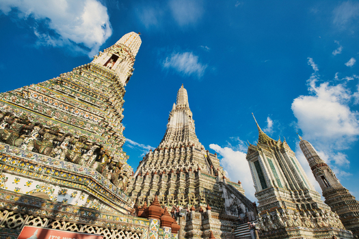 View of the ornate spires of Wat Arun temple decorated with colorful porcelain mosaics under a bright blue sky in Wat Arun Bangkok Thailand.