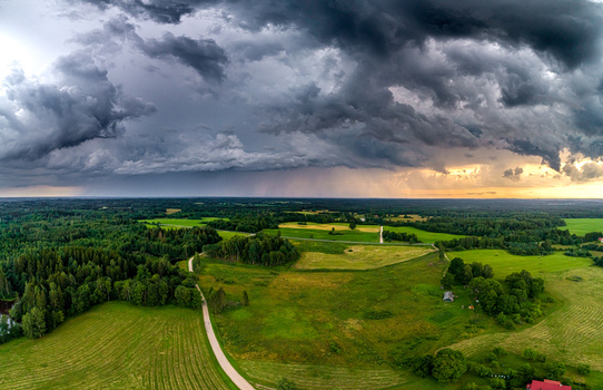 Aerial view of a rural landscape with lush green fields dense forests and a winding road under dramatic storm clouds during sunset in Latvia. Aerial view of a rural landscape with lush green fields dense forests and a winding road under dramatic storm clouds during sunset in Latvia.