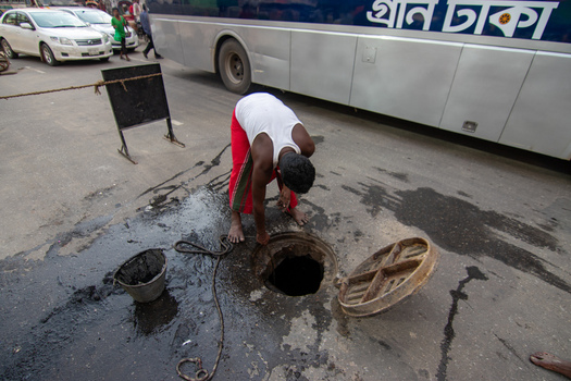 Dhaka Bangladesh 18 April 2018 View of a man cleaning a sewer through an open manhole on a busy street with a bus and cars in the background. Dhaka Bangladesh 18 April 2018 View of a man cleaning a sewer through an open manhole on a busy street with a bus and cars in the background.