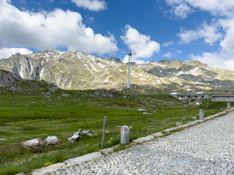 View of the historic cobblestone road at Sankt Gotthard Pass with a wind turbine and rugged mountain peaks under a cloudy sky in Airolo Ticino Switzerland.