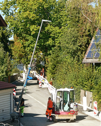 Garching Germany 28 September 2026 Aerial view of a worker using an excavator to install a new street lamp at a road construction site surrounded by trees.