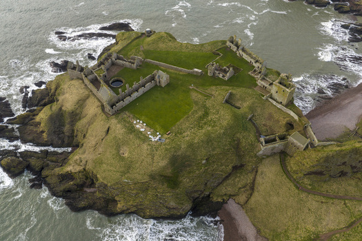 Aerial view of Dunnottar Castle ruins on a rocky cliff edge overlooking the North Sea with crashing waves and green grass Stonehaven Scotland United Kingdom. Aerial view of Dunnottar Castle ruins on a rocky cliff edge overlooking the North Sea with crashing waves and green grass Stonehaven Scotland United Kingdom.