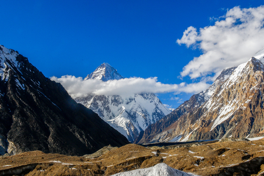 View of the snow capped peak of K2 mountain rising above rocky ridges under a clear blue sky with white clouds in Skardu Gilgit Pakistan.
