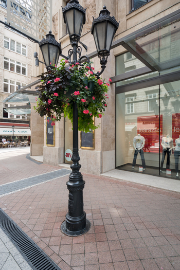 View of a decorative street lamp adorned with vibrant pink flowers on a pedestrian street in front of modern storefronts in Budapest Hungary.