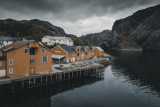 Nusfjord Norway 07 September 2022 View of Nusfjord with its iconic orange wooden buildings on stilts over the water set against dramatic rocky mountains under a cloudy sky. Nusfjord Norway 07 September 2022 View of Nusfjord with its iconic orange wooden buildings on stilts over the water set against dramatic rocky mountains under a cloudy sky.