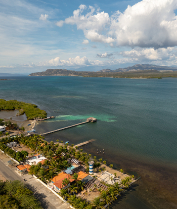 Aerial view of Las Calderas featuring a wooden pier extending into the turquoise bay a small lighthouse lush palm trees and coastal buildings in Bani Peravia Dominican Republic.