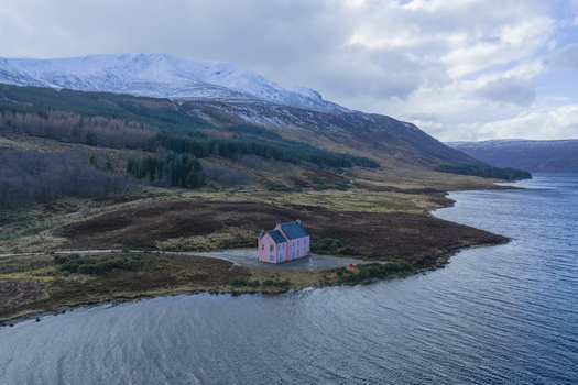 Aerial view of The Pink House on the shore of Loch Glass with snow capped mountains and a cloudy sky in Dingwall Scotland United Kingdom. Aerial view of The Pink House on the shore of Loch Glass with snow capped mountains and a cloudy sky in Dingwall Scotland United Kingdom.