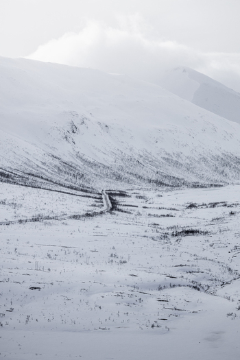 View of a winding road through a snow covered valley surrounded by majestic mountains under an overcast sky in Ersfjord Troms Norway. View of a winding road through a snow covered valley surrounded by majestic mountains under an overcast sky in Ersfjord Troms Norway.