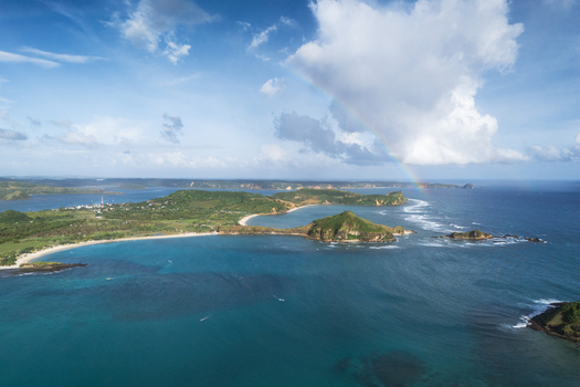 Aerial view of Tanjung Aan Beach featuring curved white sand bays turquoise waters and a faint rainbow under a cloudy sky in Kecamatan Pujut Nusa Tenggara Barat Indonesia.